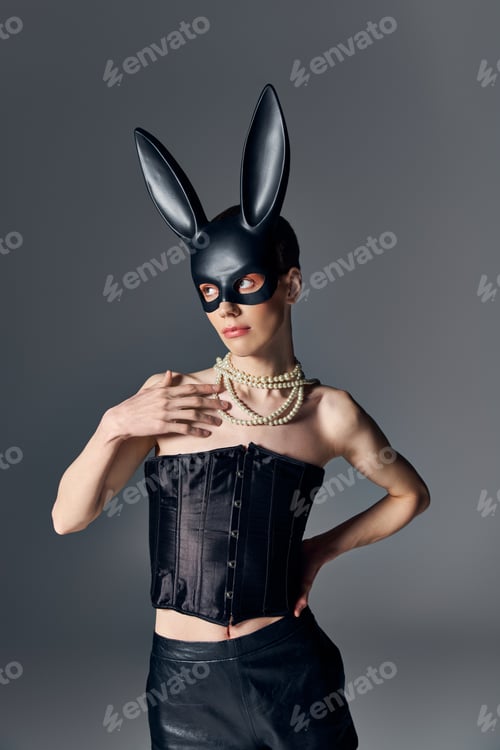 Preview: fashion, genderqueer person in corset posing in bdsm bunny mask on grey, queer style, hand on hip