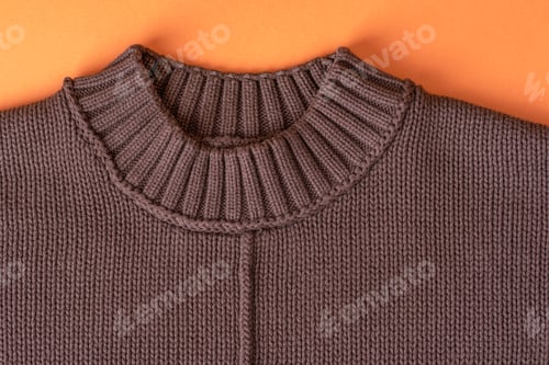 Preview: A close view of a knitted brown sweater with a ribbed collar placed on an orange background
