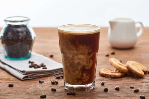 Preview: selective focus of ice coffee mixing with milk in glass near milk jug, cookies and coffee grains on