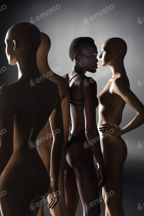 Preview: young african american model in bodysuit standing between dummies on black