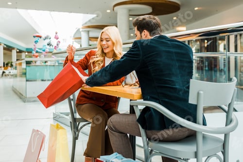 Preview: beautiful happy woman showing something in shopping bag to boyfriend while sitting in cafe in