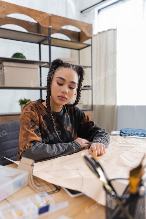 Preview: African american designer looking at sewing pattern near blurred equipment in workshop