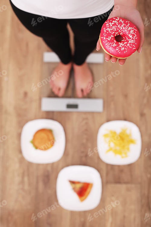 Preview: Young beautiful woman standing on scales and holding a donut.