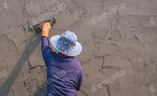 Preview: Rear view of female construction laborer using trowel to plastering cement fence wall