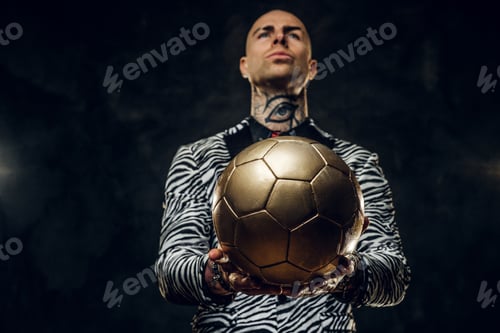 Preview: Elegant tattooed male model posing with a golden soccer ball in a studio