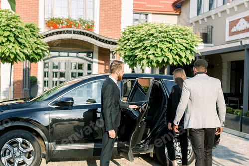 Preview: Chauffeur man opening car door for business couple, Caucasian woman and African man, side view