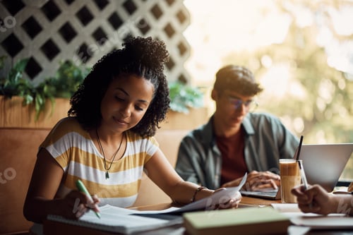 Preview: Black college student writing in notebook while preparing for exams with her friends in cafe.