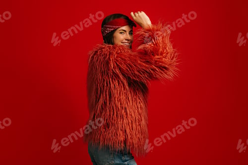 Preview: Attractive young hipster woman wearing fluffy coat standing against red background