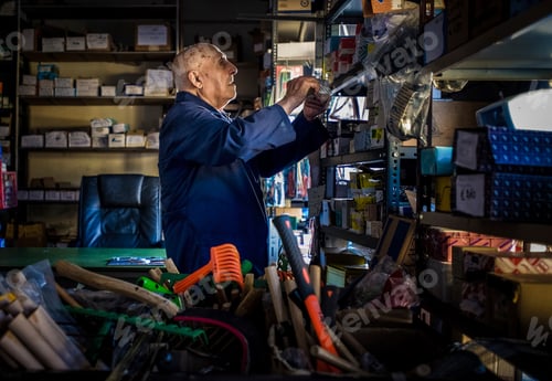 Vista previa: Elderly man meticulously arranging items on a storeroom shelf.