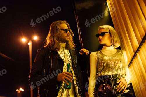 Preview: young attractive couple in stylish clothes and sunglasses flirting on night street under yellow