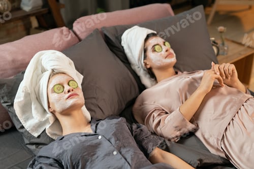 Preview: Two young women with clay masks on faces enjoying beauty procedure on bed