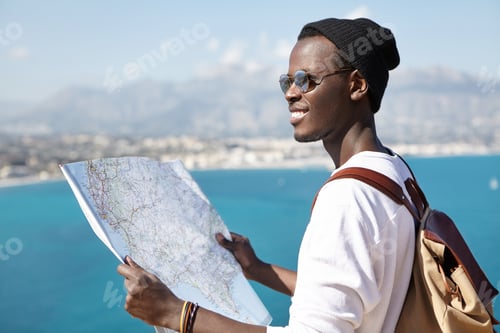 Preview: What a beautiful landscape! Happy excited Afro American backpacker using paper map while standing on