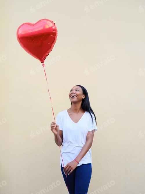 Preview: Cheerful african american woman holding a red heart balloon outdoors.