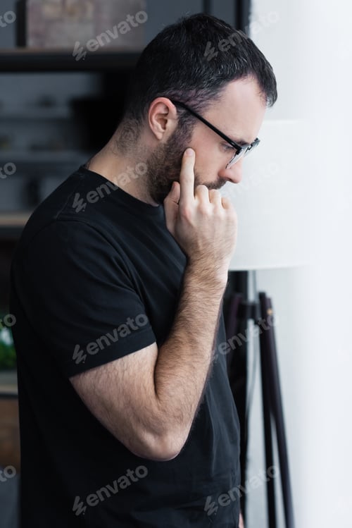 Preview: side view of upset man in black t-shirt and glasses holding hand near face