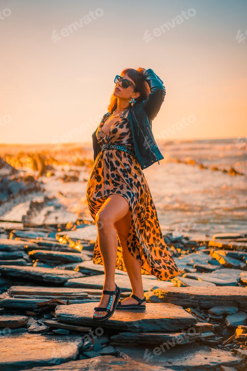 Preview: Summer street style on a beach next to rocks of a young brunette caucasian woman in a leopard dress