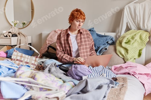 Preview: Busy redhead woman works on laptop computer sells clothes online surrounded with variety of clothing
