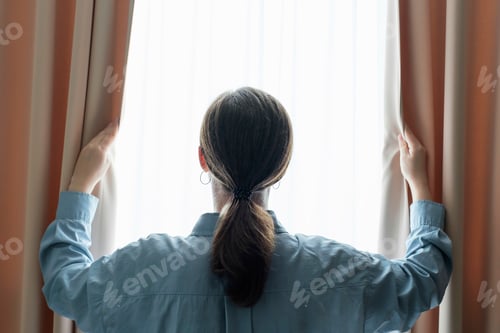 Preview: Young woman stands at window and opens curtains back view.