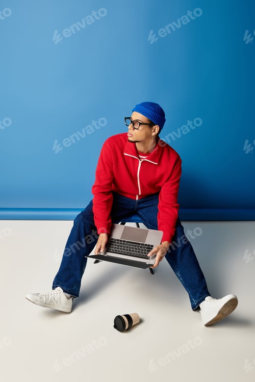 Preview: Stylish young man in beanie works on laptop beside coffee cup against blue backdrop