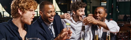 Preview: excited multiethnic friends holding tequila shots with lime near glasses of beer in bar, banner