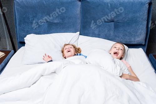 Preview: adorable happy kids having fun while lying under blanket in bed