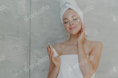 Preview: Woman holds bottle of body lotion, applies face cream on complexion, stands with closed eyes