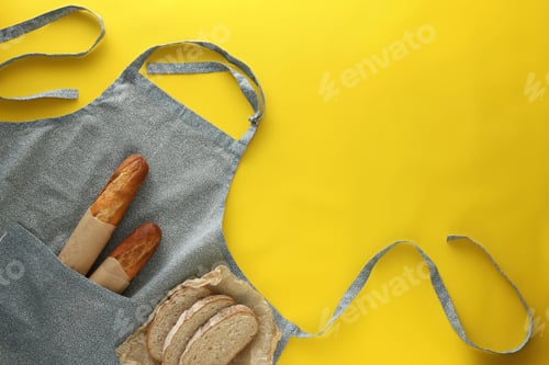 Preview: Clean kitchen apron with different types of bread on yellow background, flat lay. Space for text