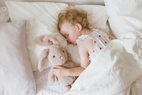 Preview: Little baby sleeping in bed with big soft pillows and white linen, hugging soft plush toy bunny.