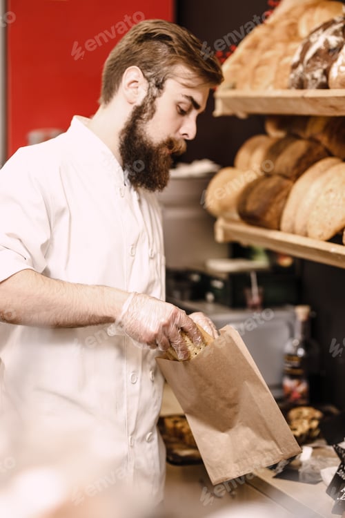 Preview: Charismatic baker with a beard and mustache puts fresh bread in a paper bag in the bakery