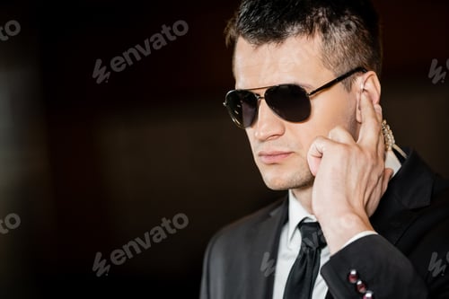 Preview: good looking bodyguard in sunglasses, handsome man in suit touching earpiece in lobby of hotel