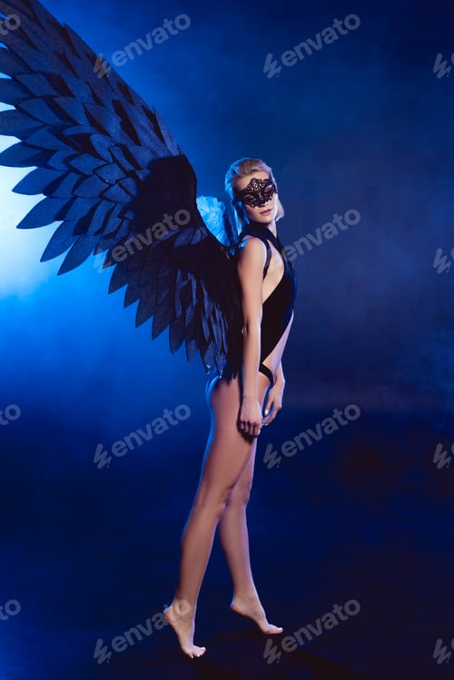 Preview: beautiful sexy woman with lace mask and black angel wings standing on tiptoe on dark blue background