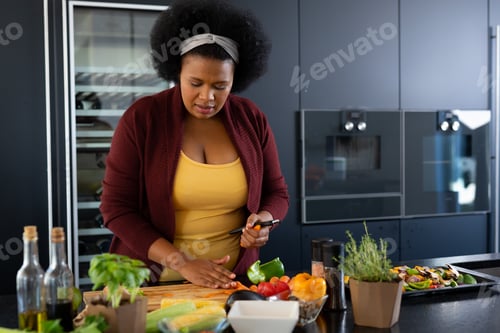 Preview: Happy plus size african american woman preparing dinner, chopping vegetables in kitchen
