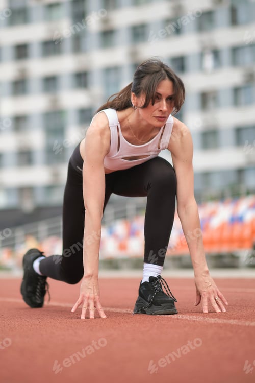 Preview: A determined woman in sportswear crouches at the starting line on a running track, ready to sprint