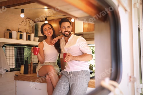 Preview: Happy young couple with cups in trailer, view from outside. Camping vacation