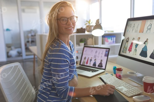 Preview: Young blonde female fashion designer looking at camera while working at desk in a modern office