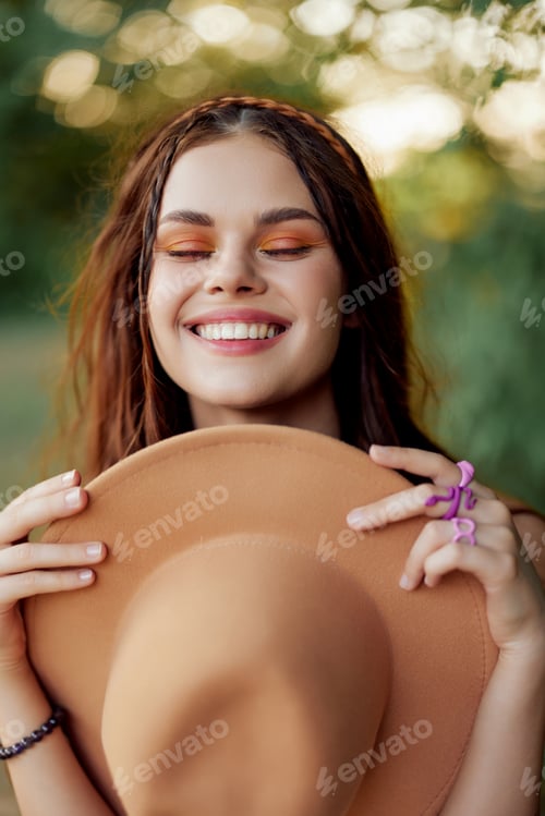 Preview: Young hippie woman holding a hat and smiling sincerely while looking at the camera in nature in the