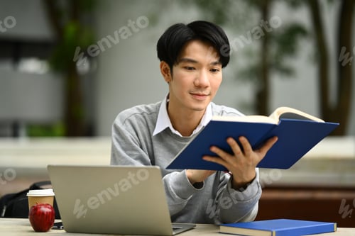 Preview: Peaceful young asian man student reading interesting book while sitting bench in sunny summer park