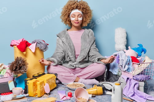 Preview: Indoor shot of relaxed curly haired young woman sits crossed legs in lotus pose dressed in domestic