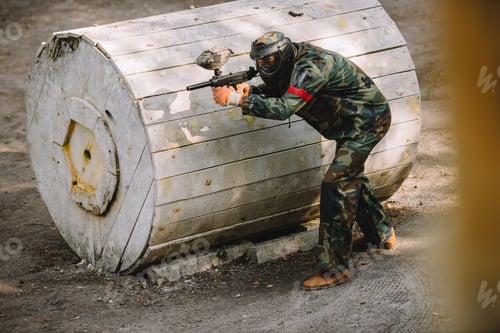 Preview: male paintball player in goggle mask and camouflage aiming by paintball gun outdoors