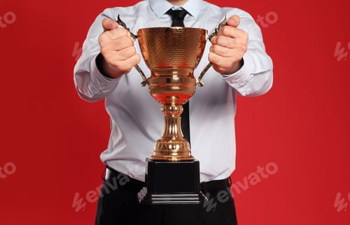Preview: Young businessman with gold trophy cup on red background, closeup