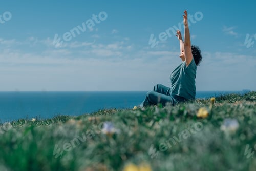 Preview: happy adult woman raises her hands to the sky in the summer in the mountains, sitting on the grass