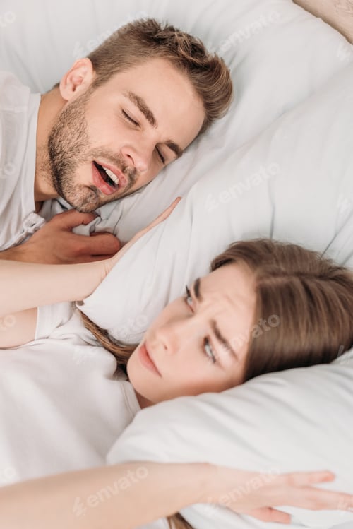 Preview: awakened woman plugging ears with pillow while lying in bed near snoring husband