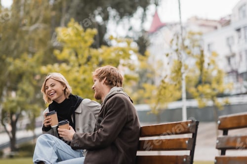 Preview: joyful young couple laughing and holding paper cups with coffee to go while sitting on bench in park