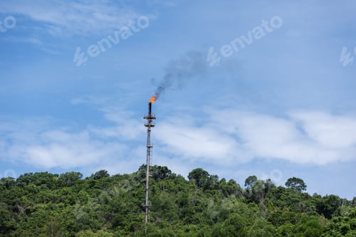 Preview: A gas flare emits smoke and flames from a tall industrial stack situated near a forested area
