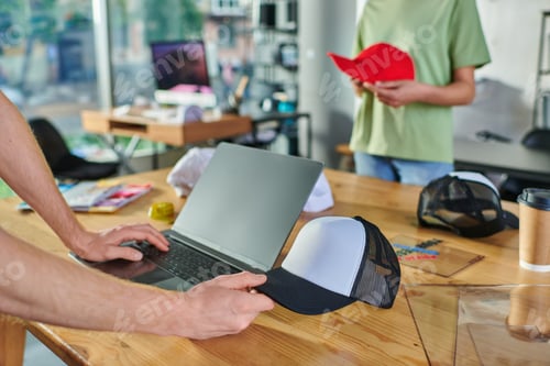 Preview: Cropped view of young designer using laptop and holding snapback and working near blurred african