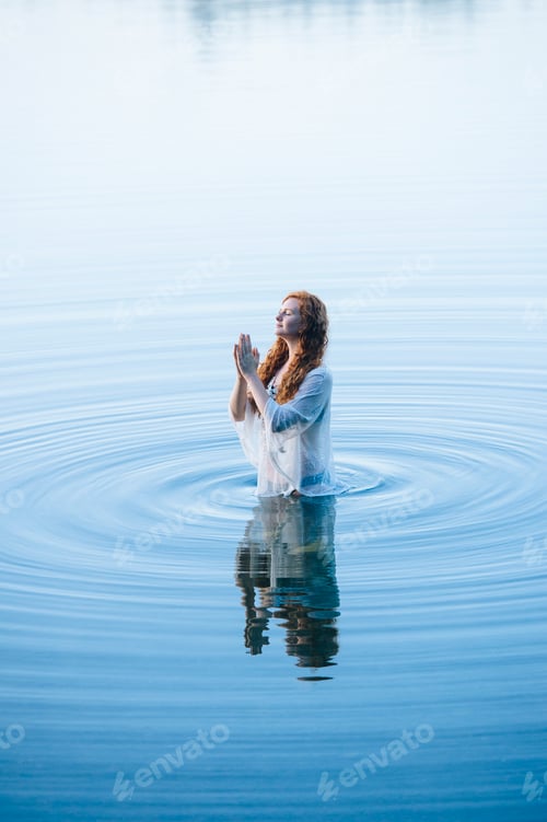 Preview: Young woman standing in lake ripples with eyes closed and hands together