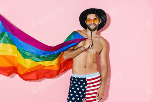 Preview: Shirtless man in hat laughing while posing with rainbow flag