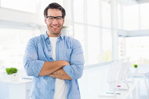 Preview: Mid-30s man standing arms-crossed, smiling, wearing glasses, striped shirt in office with monitors