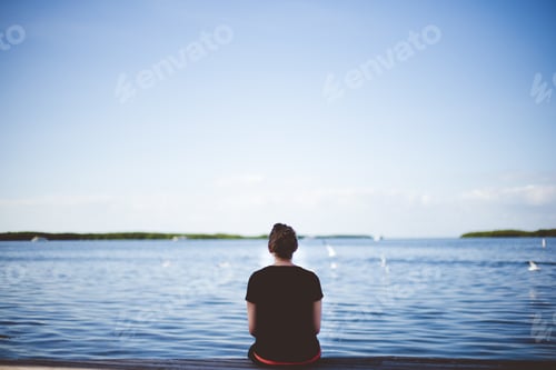 Preview: Female sitting near the sea and looking in the distance shot from behind with a blurred background
