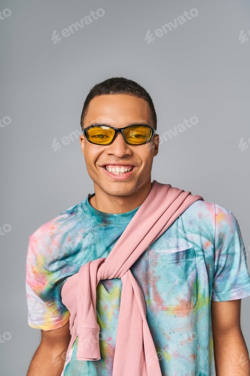 Preview: happy african american man in sunglasses and tie-dye t-shirt looking at camera on grey