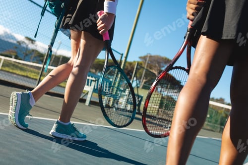 Preview: Low section of female multiracial tennis players walking rackets at court on sunny day